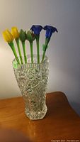 Clear 10-inch tall crystal vase filled with six long stem glass flowers including yellow tulips and blue bell-shaped flowers, standing on wood surface.