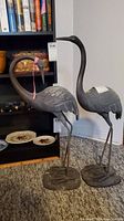 Both heron statues side by side in front of bookshelf