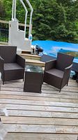 Photo of two brown resin wicker/rattan patio chairs and matching glass topped side table placed on wooden deck near pool area.