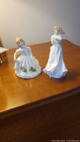Two Royal Doulton figurines on wooden surface, left is May Figure of the Month, right is Sentimental Thank You figure.