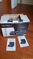 Opened box with Victrola Bluetooth turntable inside and instruction manuals placed outside the box on hardwood floor.