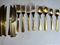 Photo showing multiple gold-tone stainless steel knives, forks, and spoons arranged on a white surface, some forks wrapped in plastic.