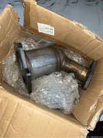 Catalytic converter shown packaged in cardboard box with bubble wrap, displaying the cylindrical metal component and flanged ends.