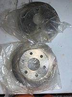 Two new metal brake rotors wrapped in clear plastic on a white surface, showing different angles of the rotors.