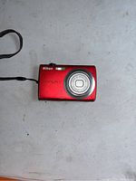 Front side of red Nikon Coolpix S203 showing lens and wrist strap on gray surface.