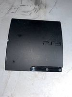 Black Sony PlayStation 3 Slim console with PS3 logo on top, shows surface wear and scratches, photo taken top-down.