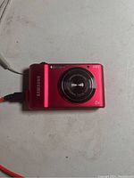 Red Samsung ST66 digital compact camera front with lens and flash visible, connected to USB charging cable on white surface.