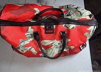 Full view of red duffel bag with dollar bill graphic print and black handles showing the tear at the top edge