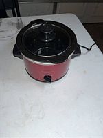 Front angled view of red Toastmaster crockpot slow cooker with black handles and glass lid, placed on a white surface.