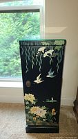 Front view of tall black lacquer cabinet with birds, flowers, ivy, and bamboo painted in multiple colors, closed door with metal ring handle.