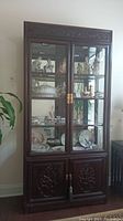 Front view of tall solid wood China cabinet with glass doors showing interior china and figurines, carved motifs on lower doors.