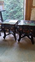 Pair of Asian design carved wood end tables with painted bird and floral designs on top and sides, set on a concrete floor by a window showing natural light and greenery outside.
