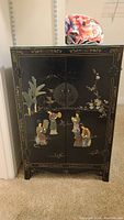 Front view of vintage black Asian cabinet with inlaid hardstone figures and hand-painted gold details.