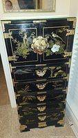 Front view of tall black jewelry valet cabinet with brass fittings and hand painted floral and bird designs on door and drawers.