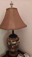 Full view of the Asian ceramic table lamp with beige shade sitting on a wooden surface.