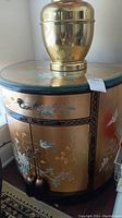 Front and side view of the curved gold and black nightstand with painted floral and bird motifs. Shows drawer, cabinet doors with tassels, and glass top with some dust/debris. A brass or gold urn is placed on top.