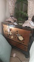 Full view of a wooden Asian-style nightstand with painted floral motifs, showing one drawer at bottom and cabinet doors, with glass top, holding decorative lamps on top and surrounded by plants.