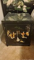 Black lacquer Asian-style cabinet with hand-painted and mother of pearl inlay scenes on front and sides, topped with glass protector and tassel on door handles.
