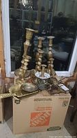 Brass candle holders of varying sizes on cardboard box in basement setting, showing tarnish and variety of styles