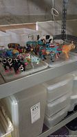 Photo showing multiple colorful cow ornaments arranged on a plastic tray on a shelf, some cows in various colors and patterns visible, with storage bins underneath.