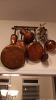 Photo showing all eight items hanging on the wooden rack including three copper frying pans, a pitcher, a ladle, a strainer, and a scoop, with visible patina and tarnish.
