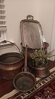 Photo showing the set of two large copper pots stacked, a small copper pot, a copper frying pan with a long handle, and a large silver plated serving tray leaning against the pots, showing patina and tarnish on all items.