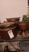 Three copper items on glass shelf: two round large pots with tarnishing and wear, one copper pedestal planter bowl. All have signs of oxidation and heavy use.