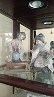 Photo showing two Lladro Geisha porcelain figurines in a cabinet, each working on floral arranging with blue vases and on stands.