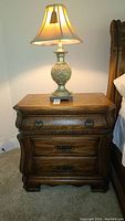 Front view of AICO oak nightstand with a lamp on top, showing ornate drawer handles and curved edges.