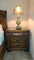 Full view of the wooden nightstand with lamp placed on top, showing its finish, shape, and size relative to bed and wall.
