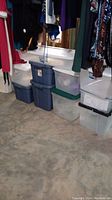 Photo of ten assorted plastic storage totes stacked in a basement closet, with clothes hanging above.