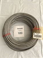 Top-down view of a tightly wound heavy metal wire cable coil placed on a white cloth background with a MaxSold card labeled 189.