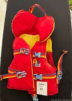 Front view of children's red and yellow life vest with rainbow-colored adjustable straps and blue plastic buckles, and overhead handle.