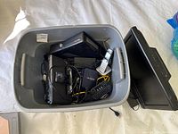 Top view of grey bin containing Xbox 360 console, wireless controller, silver cordless phone remote, black MyG Pro device, and assorted cords.