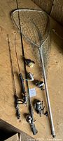 Photo shows three fishing rods each with a spinning reel attached, three additional spinning reels, and one large fishing net with an aluminum frame.
