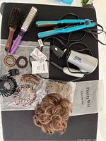 Top view of hair care items including blue flat iron, white blow dryer, two hairbrushes, several hair clips, spiral hair ties, and elastic messy bun hairpieces in bag