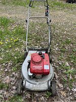 Front view of the entire lawn mower showing the red Honda HR215 engine cover and silver metal deck, standing on grassy and leafy ground.