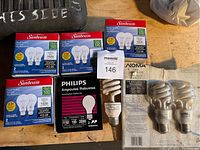 Photo showing 6 Sunbeam 100 watt rough service light bulb boxes (2 bulbs each), one Philips bulb box, one Noma spiral CFL bulb, and loose globe bulbs in Noma packaging on wood surface.