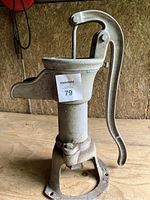 Front-left angled view showing the large curved handle, spout, and round base of the cast iron hand water pump.