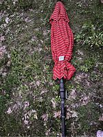 Photo of folded red patio umbrella with thin white horizontal stripes on a black pole resting on grass.
