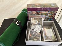 Three sealed Thomas Kinkade puzzle bags inside Thomas Kinkade puzzle box next to green puzzle mat.