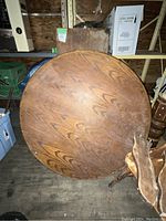 Round wooden table top in a storage area, showing wood grain and signs of sanding and wear.