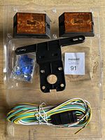 Set of two amber rectangular tail lights, black mounting bracket, wiring harness, and bag of small connectors and screws in plastic packaging.