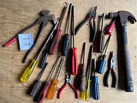 Top-down view of assorted hand tools on a wooden surface. Includes hammers, pliers, and multiple screwdrivers with plastic and wooden handles, some with red grips.