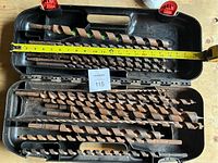 Set of large wood drill bits in black plastic case with a tape measure showing length.
