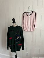 Two sweaters displayed on a mannequin and hanger on a blank wall: dark green knit with colorful design on mannequin, pink zip-up on hanger.