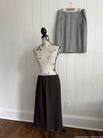 Two skirts displayed, a brown long skirt on a dress form and a gray knee-length skirt hanging on the wall.