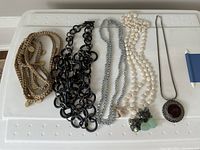 Five necklaces arranged flat showing details and variety, including the braided gold-tone rope, black link chain, two pearl strands and silver-tone pendant.