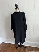 Full view of black long sleeve Ewanika dress displayed on a mannequin standing on a wooden floor against white wall.
