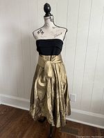 Full view of the dress on a mannequin showing the strapless black bodice and gold pleated skirt with wide gold sash tied at the waist.